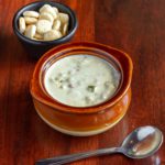 a bowl of clam chowder framed with a soup spoon and a cup of oyster crackers.