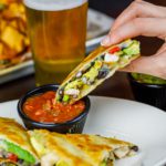 A hand holding 1 of 4 quesadilla triangles is dipping the tip into salsa. In the background sits a beer and a platter.