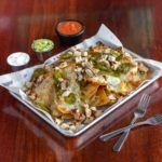 A large baking sheet piled with nacho chips covered with cheese, jalapenos, and chicken. There sits side cups of salsa, sour cream, and guacamole. Two forks sit crossed over each other to the right.