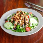 a white bowl filled with a greek salad topped with sliced chicken breast. The background has a set of silverware
